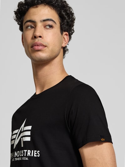 Mężczyzna model nosi czarne T-Shirt mit Label-Print od Alpha Industries z białym nadrukiem, bawełna, prosty krój, półprofil, zbliżenie na górną część ciała.