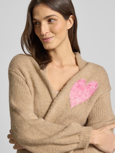 Frauenmodel trägt Longcardigan mit Eingrifftaschen von miss goodlife, Baumwoll-Alpaka-Mix, beige mit pinkem Herz, lockerer Schnitt, Nahaufnahme Oberkörper.