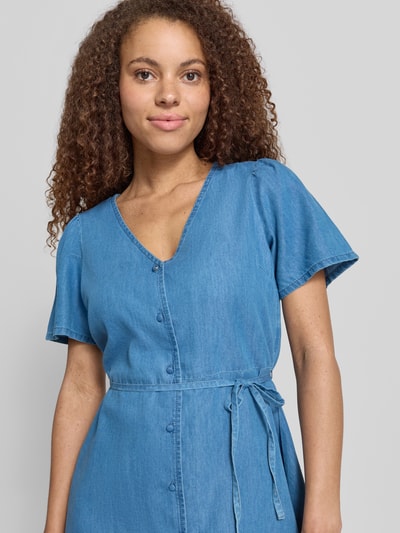 Frauenmodel trägt tailliertes Denimkleid aus Baumwoll-Lyocell-Mix mit Knopfleiste und Taillengürtel Modell 'EVIDA-BEA' von Only, Denim-Optik, Mini-Länge, V-Ausschnitt, Kurzarm, glatte Textur, Nahaufnahme Oberkörper.