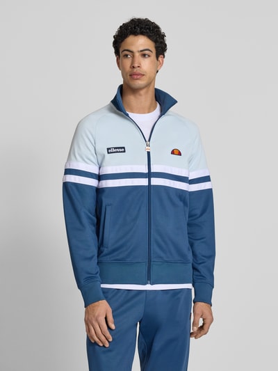 Męski model nosi bluza rozpinana z mieszanki bawełny model 'RIMINI' od Ellesse, prosty krój, stójka, zamek błyskawiczny, detale z logo, elastyczne wykończenia, długość tyłu 71 cm w rozmiarze M, długość rękawów 43 cm w rozmiarze M, odcienie niebieskiego, biel, gładka tekstura, całkowity widok z przodu.