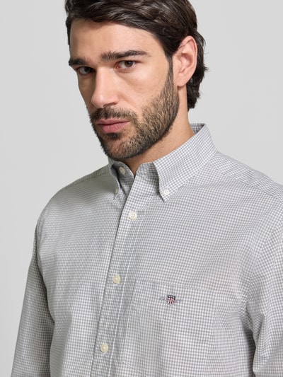 Męski model nosi Slim Fit Freizeithemd mit Logo-Stitching od Gant, szara krata, bawełna, kołnierz button-down, długi rękaw, kieszeń na piersi, szerokość w rozmiarze M: 48 cm, długość pleców: 79 cm, długość rękawa: 68 cm, zbliżenie na górną część ciała.