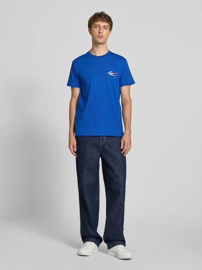 Männermodel trägt Slim Fit T-Shirt aus reiner Baumwolle von Tommy Jeans in Blau, kombiniert mit dunkler Jeans, Ganzkörperansicht von vorne.