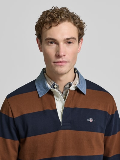 Męski model nosi Bluza z wyhaftowanym logo Sweatshirt mit Label-Stitching od Gant z bawełny, krój regular fit, kołnierzyk polo, prążkowane zakończenia, pasy w kolorze granatowym i brązowym, długość tyłu 72 cm, długość rękawów 66 cm. Nahaufnahme Oberkörper.