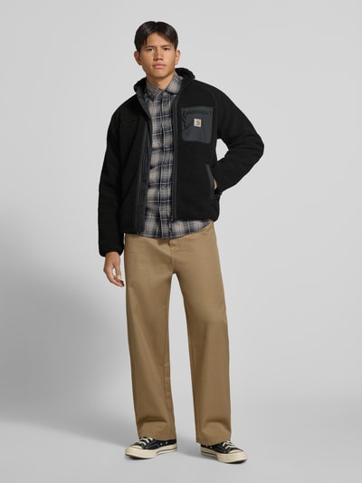 Männermodel trägt Teddyjacke mit Brusttasche Modell 'PRENTIS LINER' von Carhartt Work In Progress, schwarz, Stehkragen, Reißverschlüsse, Brusttasche, Label-Patch, Regular Fit, Kombi mit kariertem Hemd und beiger Hose, Ganzkörperansicht von vorne.