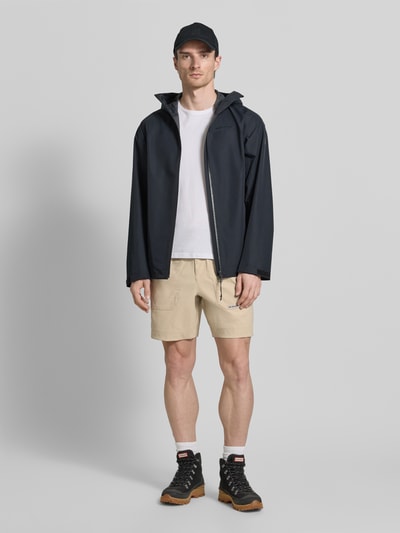 Männermodel trägt beigefarbene Regular Fit Shorts mit Logo-Patch Modell 'Landroamer' von Columbia, mit aufgesetzten Taschen und elastischem Bund, kombiniert mit Outdoor-Kleidung, Ganzkörperansicht von vorne.