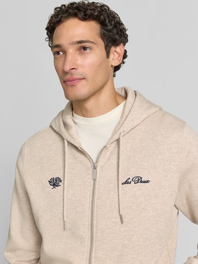 Männermodel trägt Regular Fit Sweatjacke aus reiner Baumwolle mit Logo-Stickerei von Les Deux, beige, sichtbare Teilansicht, Nahaufnahme Oberkörper. Herren Sweatjacke, Kapuze mit Tunnelzug, geteilte Kängurutasche, gerippte Abschlüsse, langärmliges Design.