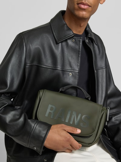 Männermodel trägt kosmetyczka Kulturtasche mit Label-Print Modell 'Texel Wash' von Rains w kolorze oliwkowym, Główna przegroda zapinany na zamek błyskawiczny dookoła, przegródki i siateczka w środku, Szerokość w rozmiarze 1: 26 cm, Długość w rozmiarze 1: 18 cm, Głębokość w rozmiarze 1: 12 cm, długość paska: 19 cm, Ganzkörperansicht von vorne