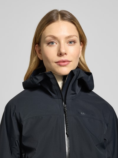 Frauenmodel trägt Mantel mit Zweiwege-Reißverschluss und Kapuze von PEAK PERFORMANCE in Schwarz, glatte Struktur, Nahaufnahme Oberkörper. Modell TREELINE HIPE® 2.5L SHELL, Relaxed Fit, Logo sichtbar.