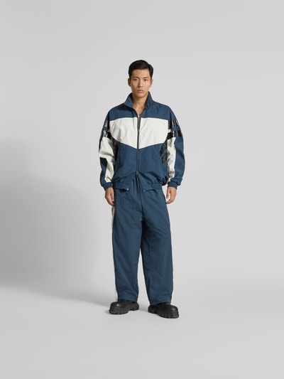 Männermodel trägt Trackpants mit elastischem Bund von VETEMENTS in Dunkelblau mit weißen Galonstreifen, Regular-Fit, Eingrifftaschen, Mesh-Futter, Ganzkörperansicht von vorne. Innenbeinlänge bei Größe M 69 cm, Bundweite bei Größe M 44 cm.