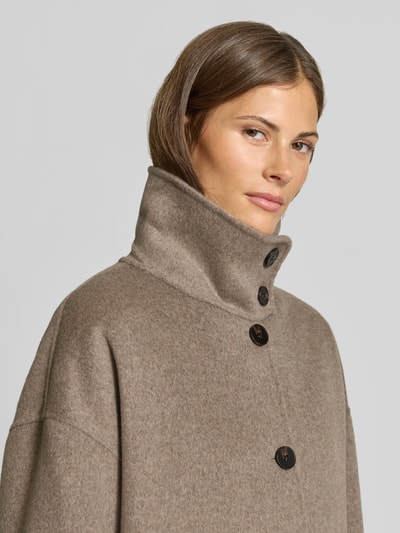 Frauenmodel trägt Wollmantel aus Woll-Mix von MYON in Beige mit hohem Stehkragen und Knopfleiste, Nahaufnahme Oberkörper. Lockere Passform, aufgesetzte Eingrifftaschen, Rückenlänge 115 cm, Schulterbreite 63 cm, Brustweite 65 cm, Ärmellänge 53 cm.