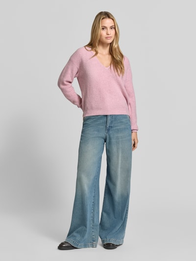 Kobieta nosi sweter z dzianiny z mieszanką lnu, Model 'DAMSVILLE' od American Vintage, efekt prążkowania, dekolt w serek, luźny krój, elastyczne wykończenia, różowy, długość tyłu 61 cm, długość rękawa 54 cm, widok całej sylwetki z przodu.