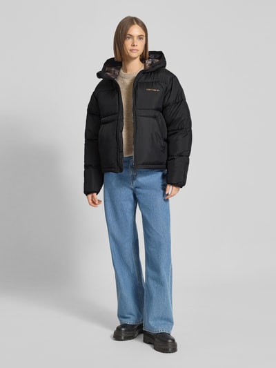 Frauenmodel trägt Steppjacke mit Kapuze und Eingrifftaschen Modell 'Toronto' von Carhartt Work In Progress, czarny kolor, krój regular fit, zamek błyskawiczny, logo, kieszenie boczne, szerokość w rozmiarze S: 60 cm, długość tyłu w rozmiarze S: 62 cm, szerokość ramion w rozmiarze S: 50 cm, długość rękawa w rozmiarze S: 62 cm, gładka tekstura, całkowity widok z przodu