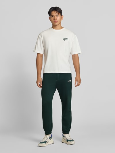 Männermodel trägt Straight Fit Sweatpants aus Baumwoll-Mix Modell 'Kane' von JACK & JONES in Dunkelgrün, Logo-Detail, Ganzkörperansicht von vorne. Herren Sweatpants aus Baumwoll-Mix mit Gesäßtasche, elastischem Bund und Beinabschlüssen, Innenbeinlänge 72 cm bei Größe M.