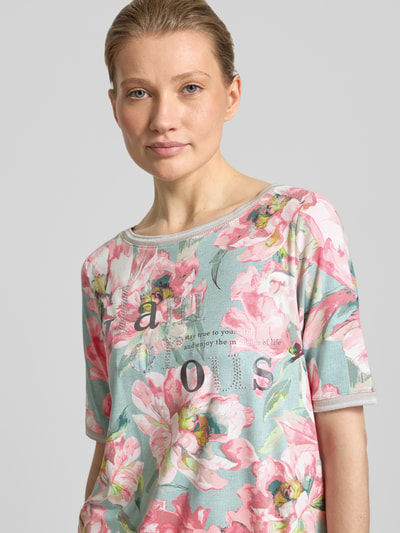Modelka kobieca nosi T-Shirt aus Viskose-Elasthan-Mix mit Statement-Print od Betty Barclay, krój regular fit, kwiatowy wzór allover, okrągły dekolt, tunel przy dole, długość tyłu 58 cm w rozmiarze 36, pastelowe kolory, tekstura miękka, zbliżenie na górną część sylwetki.