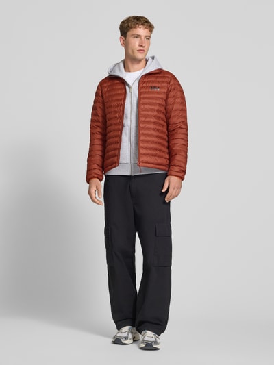 Männermodel trägt Steppjacke mit Label-Print von Helly Hansen in Braun, gerade Passform, Stehkragen, Ganzkörperansicht von vorne.