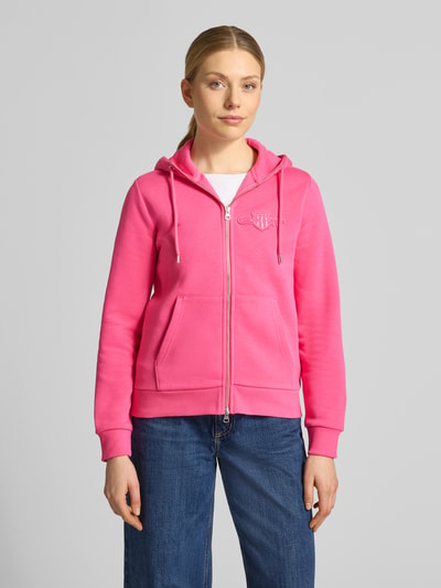 Frauenmodel trägt Sweatjacke mit Zweiwege-Reißverschluss und Label-Patch von Gant, Baumwoll-Elasthan-Mix, Regular-Fit, Kapuze, Känguru-Tasche, kräftiges Pink, strukturierte Oberfläche, Ganzkörperansicht von vorne.