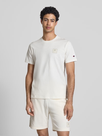 Männermodel trägt T-Shirt mit Logo und Rundhalsausschnitt von CHAMPION in Cremeweiß, reine Baumwolle, Regular Fit, Ganzkörperansicht von vorne.