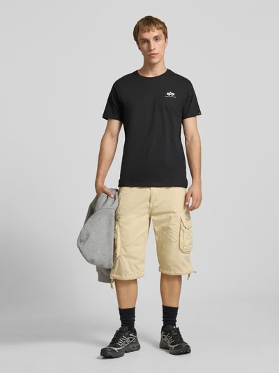 Männermodel trägt schwarzes T-Shirt mit Logo und Rundhalsausschnitt von Alpha Industries aus Reine Baumwolle, Regular-Fit, Ganzkörperansicht von vorne. Rückenlänge bei Größe M: 67 cm.
