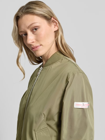 Frauenmodel trägt Bomberjacke mit Label-Patches Modell 'BROOKLYN NEO' von Frieda & Freddies in Olivgrün, strukturierte Oberfläche, Nahaufnahme Oberkörper. Gerader-Schnitt, kurzer Stehkragen, Zweiwege-Reißverschluss, Seitliche Pattentaschen mit Druckknopfverschluss, Gerippte-Abschlüsse, Rückenlänge 49 cm, Schulterbreite 42 cm, Brustweite 55 cm, Ärmellänge 63 cm, Größe 36.