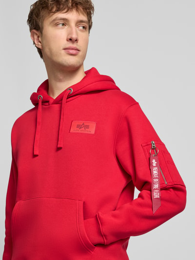 Männermodel trägt Hoodie mit Känguru-Tasche von Alpha Industries in Rot, Baumwoll-Mix, Regular-Fit, Label-Patch, Nahaufnahme Oberkörper.