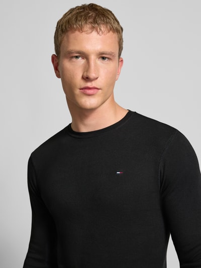 Männermodel draagt Slim Fit Longsleeve met ribstructuur van Tommy Jeans in zwart, puur katoen, geborduurd logo, ronde hals, nahaufnahme oberkörper.