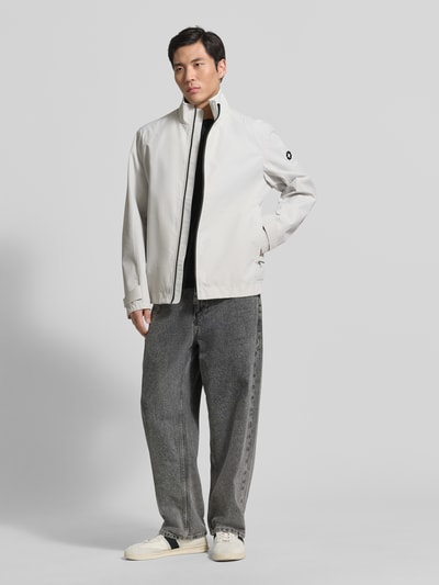 Męski model nosi Regular Fit Funktionsjacke aus wasserdichtem Material Modell 'LUCCA' von Strellson w jednolitym jasnoszarym kolorze, kieszenie na zamek, logo z metalu, kołnierz stójka, długi rękaw, tekstura gładka, krój regular fit, całkowity widok z przodu.