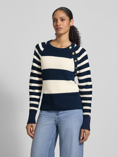 Frauenmodel trägt Regular Fit Strickpullover aus reiner Baumwolle von Tommy Hilfiger, Optik: Allover-Muster, gestreift, Zierknöpfe, Logo-Applikation aus Metall, Crew Neck, langarm mit Raglanärmel, gerippte Abschlüsse, Strick, Ganzkörperansicht von vorne.