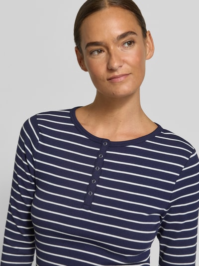 Modelka kobieca nosi Slim Fit Langarmshirt aus Baumwoll-Mix mit kurzer Knopfleiste od Christian Berg Woman, elastyczna bawełniana mieszanka, krój slim fit, okrągły dekolt, krótka listwa guzikowa, długi rękaw, długość tyłu w rozmiarze S: 58 cm, ciemnoniebieskie i białe paski, faktura prążkowana, zbliżenie górnej części ciała.