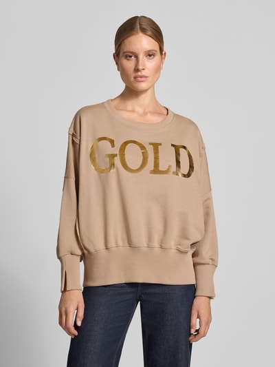 Frauenmodel trägt Oversized Sweatshirt aus reiner Baumwolle mit Statement-Print von miss goodlife in Beige mit goldfarbenem Schriftzug, Rundhalsausschnitt und elastischen Abschlüssen, Ganzkörperansicht von vorne.