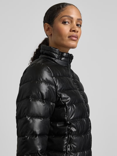 Frauenmodel trägt Jacke mit Kapuze von NAUTGÅRD in Schwarz, leicht glänzend, mit Reißverschluss und Kapuze, Nahaufnahme Oberkörper. Rückenlänge 90 cm, lockerer Schnitt.
