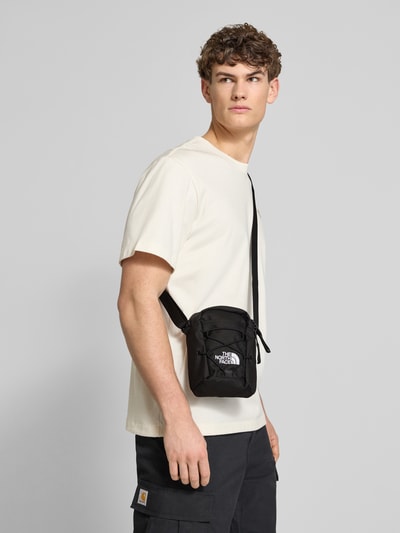 Männermodel trägt schwarze Handtasche mit Label-Stitching von The North Face, Reißverschlussfächer, Schulterriemen, Nahaufnahme Oberkörper.