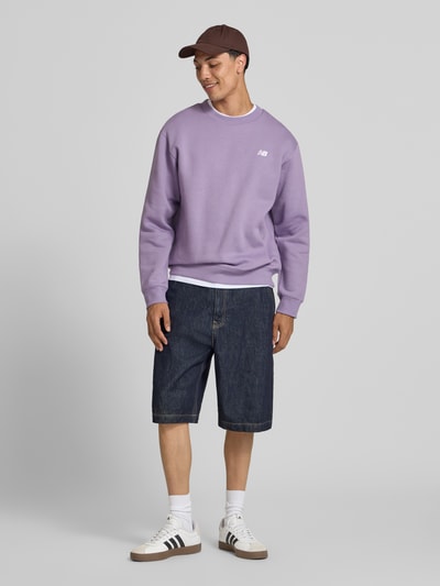 Männermodel trägt Sweatshirt mit Rundhalsausschnitt und Label-Stitching von New Balance in Lila, Baumwoll-Mix, Relaxed-Fit, Ganzkörperansicht von vorne.
