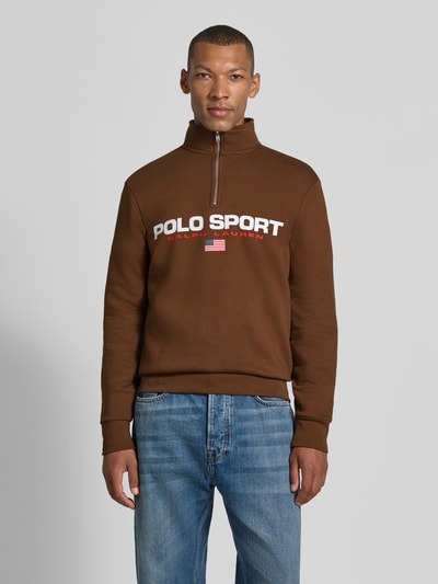 Mannelijk model draagt Relaxed Fit Troyer aus Baumwoll-Mix van Polo Ralph Lauren in bruin met witte en rode letters, blauwe jeans, Ganzkörperansicht van voren. Heren schipperstrui.