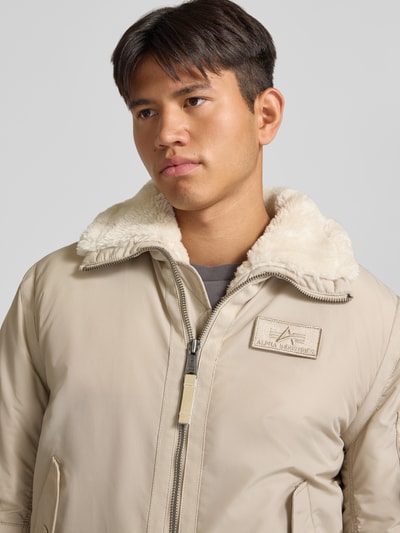 Männermodel trägt Bomberjacke mit Label-Patch und Kentkragen von Alpha Industries in Beige, sichtbarer Teddyfellkragen, Nahaufnahme Oberkörper. Regular Fit, Reißverschluss, Brustweite 56 cm, KI-modifiziertes Bild.