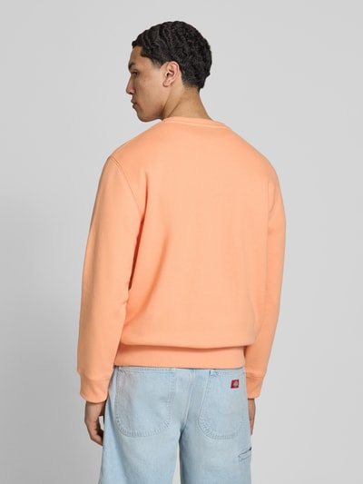 Männermodel trägt Sweatshirt mit Rundhalsausschnitt und Label-Stitching von Superdry in Pfirsich-Orange, Baumwoll-Mix, Regular-Fit, Rückenlänge bei Größe M 61 cm, Ärmellänge bei Größe M 64 cm, glatte Textur, Ganzkörperansicht Rückansicht.