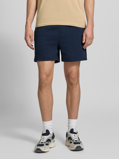 Männermodel trägt Regular Fit Shorts aus Baumwoll-Mix von Tommy Jeans, efekt: jednolity kolor, detal z logo, elastyczny tunel, kieszeń z tyłu, wpuszczane kieszenie, wykończenie: dzianina dresowa, materiał: mieszanka bawełny, całkowity widok z przodu.