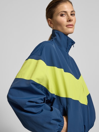 Vrouwenmodel draagt Nylon-Jacke im 90s-Stil - curated by Tinvcb van Tinvcb in blauw en geel, regular fit, staande kraag, ritssluiting, elastische boorden, steekzakken met ritssluiting, ruglengte 60 cm, schouderbreedte 54 cm, borstwijdte 58 cm, mouwlengte 34 cm, close-up bovenlichaam.