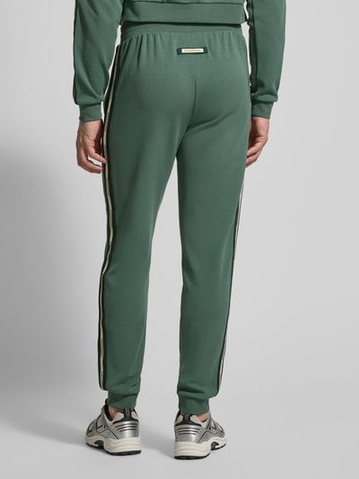 Männermodel trägt Sweatpants aus Baumwoll-Mix von EA7 Emporio Armani in Grün mit schwarzen Streifen, elastischem Bund, Eingrifftaschen, lockere Passform, Rückansicht Ganzkörperansicht.
