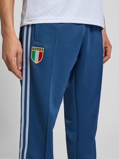 Männermodel trägt Regular Fit Sweatpants mit katoenmix model 'FIGC OG' van adidas Originals, look: effen kleuren, applicatie met labelstitching, contraststrepen, sluiting: tunnelkoord, boord: elastisch, materiaalverwerking: sweat, materiaal: materiaalmix met katoen, nahaufnahme onderlichaam.