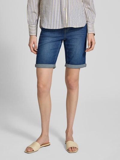 Kobiece model prezentuje szorty jeansowe z brzegiem i elastycznym pasem Jeansshorts mit Umschlag und elastischem Bund od Angels, mieszanka bawełny i elastanu, krój regular fit, szerokość w rozmiarze 36: 39 cm, długość wewnętrzna nogawki w rozmiarze 36: 30 cm, niebieski denim, całkowity widok z przodu.