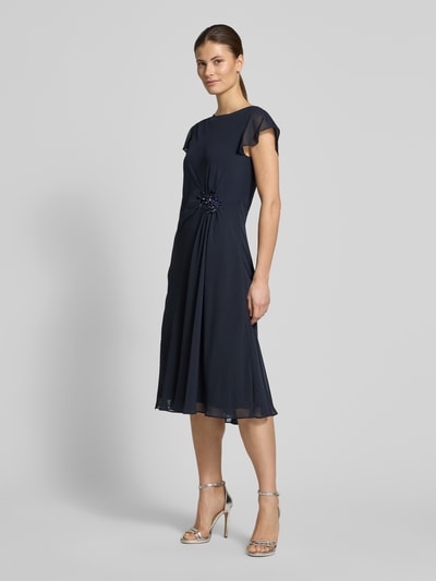 Frauenmodel trägt knielanges Cocktailkleid mit Ziersteinbesatz von Christian Berg Cocktail aus dunkelblauem Chiffon, tailliert geschnitten, Rundhalsausschnitt, kurze Ärmel, fließende Textur, Ganzkörperansicht von vorne.