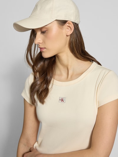 Modelka nosi Cropped T-Shirt in Ripp-Optik mit Label-Badge od Calvin Klein Jeans, bawełna z elastanem, kolor jasny beż, faktura prążkowana, badge na piersi, krótka długość, długość pleców w rozmiarze S: 36 cm, zbliżenie na górną część ciała.