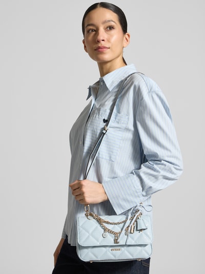 Kobieta modelka nosi torbę na ramię Umhängetasche mit Label-Applikation Modell 'LIBBY' od Guess, jasnoniebieska, aplikacja z metką, łańcuszki, główna przegroda z zapięciem na magnes, szerokość w rozmiarze 1: 24 cm, długość w rozmiarze 1: 14 cm, głębokość w rozmiarze 1: 6 cm, długość paska na ramię w rozmiarze 1: 114 cm, widok całej sylwetki z przodu.