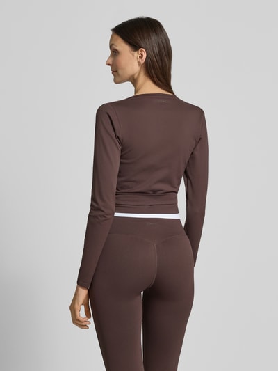 Modelka nosi Cropped Wickelshirt mit V-Ausschnitt von aim'n, mieszanka materiałów z dodatkiem streczu, krój krótki, bluzka z długim rękawem, dekolt w serek, długość tyłu w rozmiarze S: 40 cm, ciemnobrązowy kolor, gładka faktura, widok całej sylwetki z tyłu.
