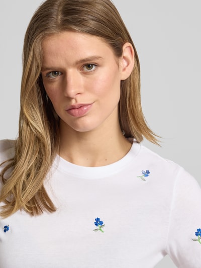 Vrouwenmodel draagt Regular Fit T-Shirt uit pure biokatoen model 'KETTY LIFE' van Only, effen wit met blauw geborduurde bloemen, ronde hals, korte mouw, detailopname bovenlichaam.