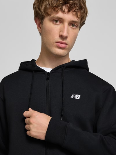 Männermodel trägt schwarze Sweatjacke mit Känguru-Tasche und Label-Stitching von New Balance, Baumwoll-Mix, Nahaufnahme Oberkörper.
