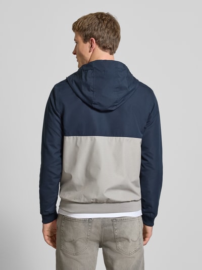 Mannelijk model draagt Jacke met capuchon en ritssluiting model 'RUSH' van JACK & JONES, regular fit, ruglengte maat M 68 cm, schouderbreedte 46 cm, borstwijdte 55 cm, mouwlengte 68 cm, kleur donkerblauw en grijs, met steekzakken en logo, geheel van achteren.