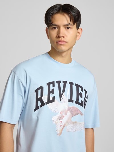 Mężczyzna model nosi T-Shirt mit Logo- und Motiv-Print od REVIEW, jasnoniebieski, czarna typografia, bawełna, krój loose fit, zbliżenie na górną część ciała.
