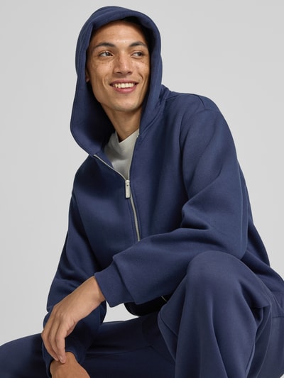 Männermodel trägt Sweatjacke aus Baumwoll-Mix von REVIEW in Dunkelblau, lockere Passform, Nahaufnahme Oberkörper, Context: Kängurutasche, gerippte Abschlüsse, Rückenlänge 70 cm, Ärmellänge 63 cm.