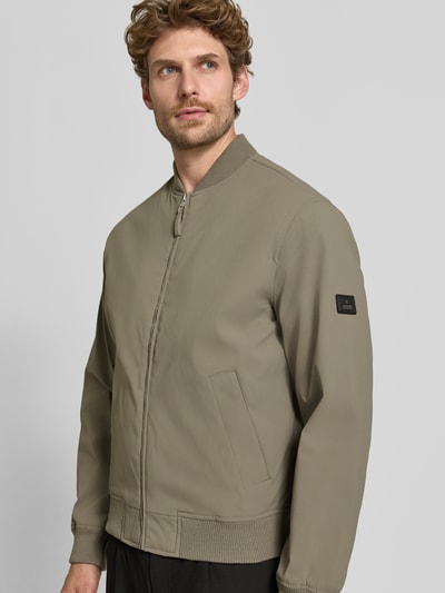 Męski model nosi bomberkę z zamkiem błyskawicznym i wpuszczanymi kieszeniami Bomberjacke mit Reißverschluss und Eingrifftaschen von JACK & JONES premium, krój regular fit, stójka, zamek błyskawiczny, wpuszczane kieszenie, długość tyłu w rozmiarze M: 66 cm, szerokość ramion w rozmiarze M: 42 cm, szerokość klatki piersiowej w rozmiarze M: 60 cm, długość rękawów w rozmiarze M: 68 cm, kolor khaki, matowa tekstura, widok całej sylwetki z przodu.